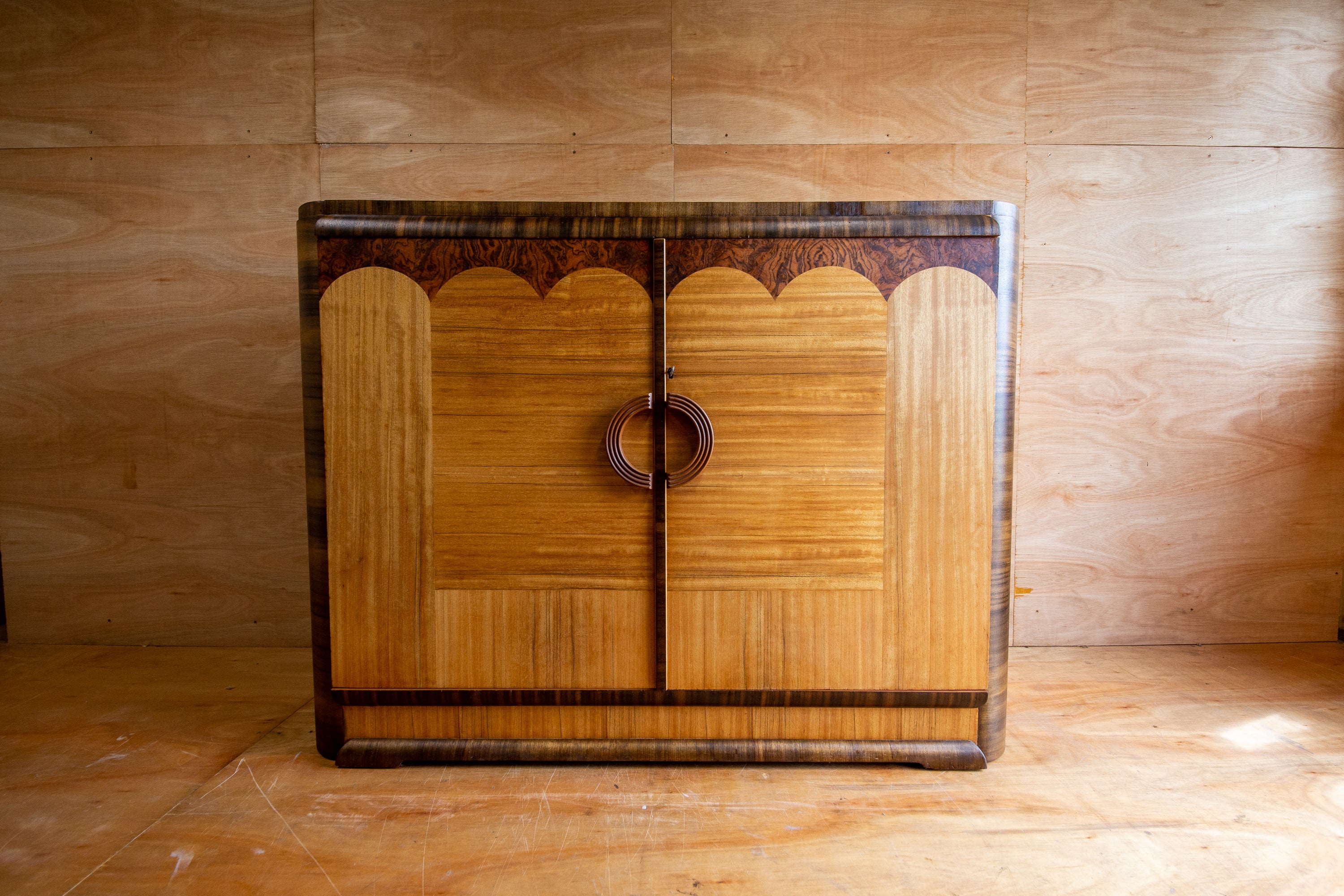 Vintage Art Deco style sideboard with brown marquetry veneer work and curved handles, featuring a frontal door with a lock and key, placed against a wooden background.
